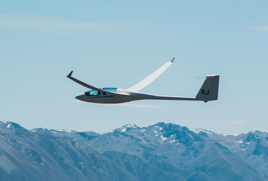 Sleek glider soaring over snow-capped mountains in Omarama, New Zealand, under a clear blue sky.